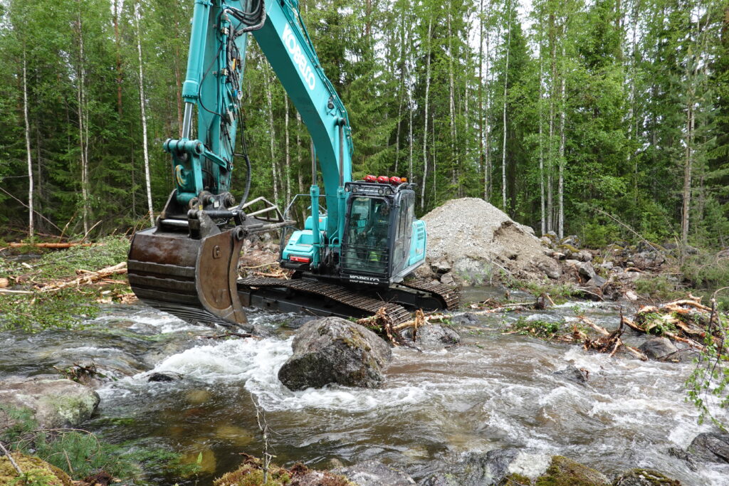 Grävmaskin i vattendrag med stora block.