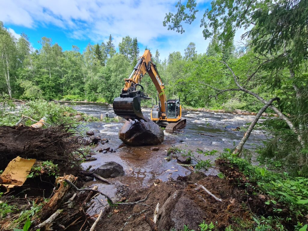 Grävmaskin som står i vatten och jobbar med ett stort stenblock.